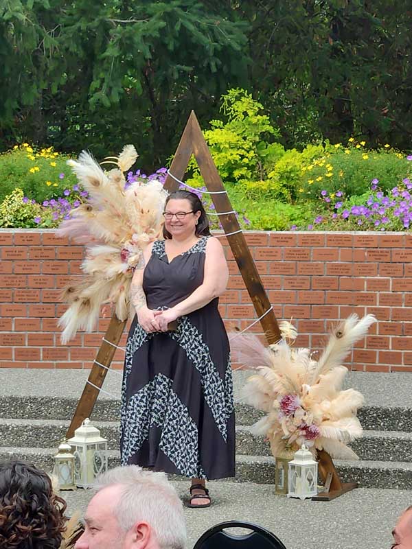 Wedding officiant standing in front of a rustic wooden triangle arch decorated with pampas grass, flowers, and lanterns, ready to lead a ceremony.